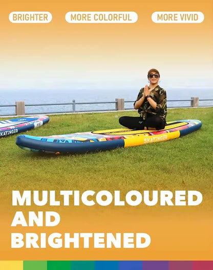 A person sits cross-legged on the Skatinger Graffiti Paddle Board on grass by the ocean, smiling with hands together. The image features a bright orange gradient border and text: “MULTICOLOURED AND BRIGHTENED.”.